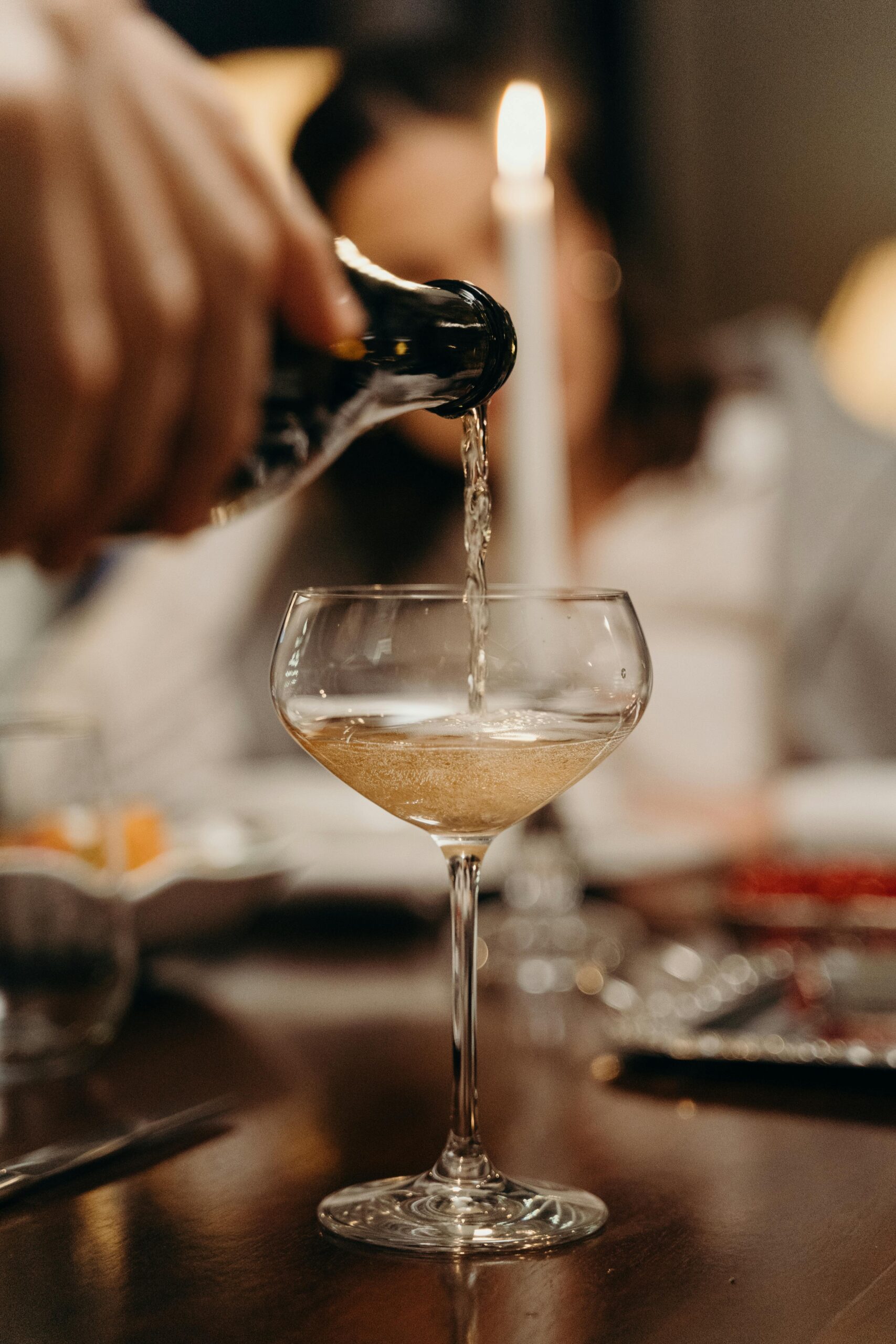 A close-up of champagne being poured into a coupe glass at a candlelit dinner setting.